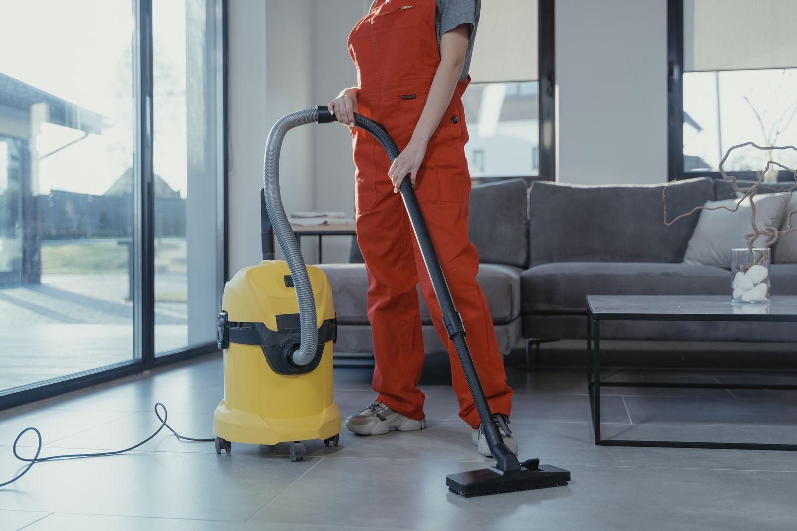 A professional cleaner in red coveralls vacuuming a modern living room with a yellow vacuum cleaner